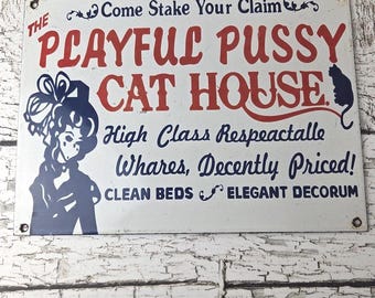 May include: Vintage metal sign with the text "THE PLAYFUL PUSSY CAT HOUSE" in red and blue lettering. The sign features a silhouette of a woman and a cat. Additional text includes "High Class Respeactalle Whares, Decently Priced! CLEAN BEDS ELEGANT DECORUM."