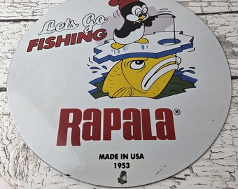 May include: Round metal sign with a cartoon penguin fishing through a hole in the ice, above a yellow fish. The sign reads "Let's Go Fishing" and "Rapala", with "Made in USA 1953" below.