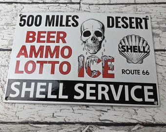 May include: A rectangular white metal sign with black and red text and graphics. The sign features a skull, the Shell logo, and the words "500 Miles", "Beer", "Ammo", "Lotto", "Desert", "Ice", "Shell Service", and "Route 66".