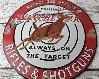 May include: A circular metal sign featuring a red and white design with a leaping deer in the center. The text reads AUTHORIZED DEALER, RIFLES BRAND WINCHESTER, ALWAYS ON THE TARGET, and RIFLES & SHOTGUNS. The sign has mounting holes on the sides.