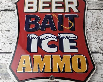 May include: A vintage metal sign with a red background and white, blue, and yellow lettering. The sign reads "BEER BAIT ICE AMMO" in bold, block letters. The sign has a shield shape with a white border and two holes for hanging.