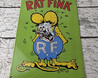 May include: A green metal sign featuring the cartoon character Rat Fink. The sign has the words "RAT FINK" in red at the top. The character is a grotesque mouse-like creature wearing blue overalls with "RF" on the front. The sign is dated '63.
