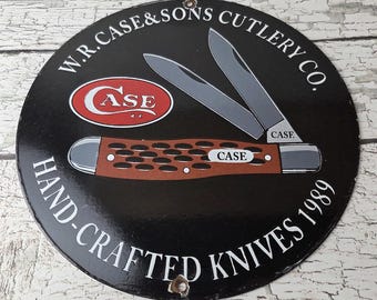 May include: A round black metal sign featuring the text "W.R. Case & Sons Cutlery Co." and "Hand-Crafted Knives 1989." The sign has a red and white "Case" logo and an illustration of a pocket knife.