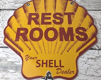 May include: Vintage Shell gas station sign in the shape of a yellow seashell with red trim and lettering. The sign reads "REST ROOMS" and "Your SHELL Dealer". The sign has two holes for hanging.