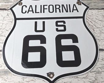 May include: A white and black metal sign in the shape of a shield, with the words "CALIFORNIA" at the top and "US 66" below. The sign has a black border and some rust spots.