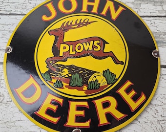May include: Round metal John Deere Plows sign. The sign features a yellow center with a brown deer leaping over a log, with the word "PLOWS" in brown. The outer ring is black with the words "JOHN DEERE" in yellow.