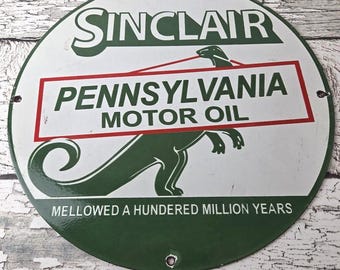 May include: Vintage Sinclair Pennsylvania Motor Oil sign. The round, white sign features green text and a green dinosaur graphic. The text includes "Sinclair" at the top, "Pennsylvania Motor Oil" in a red box, and "Mellowed a Hundred Million Years" at the bottom.