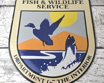 May include: Vintage U.S. Fish & Wildlife Service metal sign. The sign features a duck in flight, a leaping fish, and a yellow sun over an orange mountain range. The text "U.S. FISH & WILDLIFE SERVICE" and "DEPARTMENT OF THE INTERIOR" are displayed.