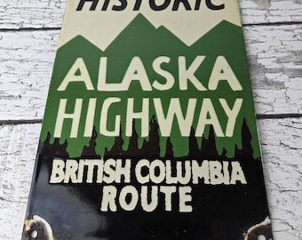 May include: A rectangular metal sign with a cream, green, and black color scheme. It features the text HISTORIC ALASKA HIGHWAY BRITISH COLUMBIA ROUTE in black and cream lettering, with a graphic of green mountains and black pine tree silhouettes.