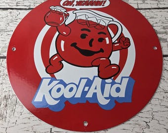 May include: A round, red metal sign featuring the Kool-Aid Man with the text "OH, YEAHHH!" in a speech bubble. The Kool-Aid Man is red with a smiling face and is holding a pitcher of Kool-Aid. The Kool-Aid logo is in blue and white.