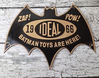 May include: A vintage Batman sign in the shape of a bat, with the words "ZAP!" and "POW!" on either side. The sign features the text "1966 IDEAL BATMAN TOYS ARE HERE!" in a black and tan color scheme.