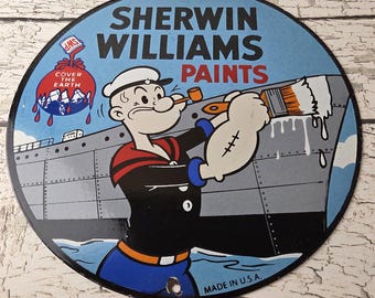 May include: Round metal sign featuring Popeye the Sailor painting a ship with white paint. The sign has a blue background with the words "SHERWIN WILLIAMS PAINTS" in bold letters. The sign also includes the text "COVER THE EARTH" and "MADE IN U.S.A."