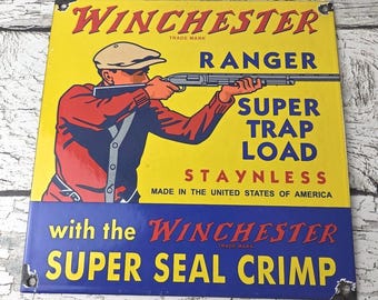 May include: Vintage Winchester Ranger Super Trap Load sign. The sign features a yellow background with a red and blue illustration of a person aiming a rifle. Text includes "Winchester", "Ranger", "Super Trap Load", and "Super Seal Crimp".