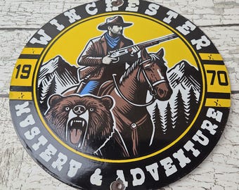 May include: A circular metal sign featuring a yellow and black design with a cowboy on horseback holding a rifle, a bear, and mountain scenery. The text reads WINCHESTER 1970 MYSTERY & ADVENTURE. The sign has two small holes for mounting purposes.