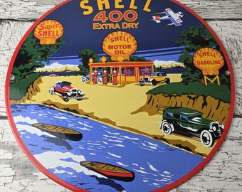 May include: A round metal sign featuring a vintage Shell 400 Extra Dry advertisement. The graphic depicts a gas station, classic cars, boats on a river, and an airplane. The text reads Shell 400 Extra Dry, Super Shell Gasoline, and Shell Motor Oil.