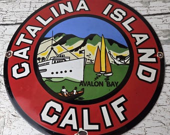 May include: A circular metal sign featuring a red border with the white text CATALINA ISLAND CALIF. The center depicts a white ship, orange and yellow sailboats, and a swimmer in blue water near green hills, with the text AVALON BAY printed below.