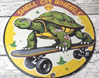 May include: A round metal sign featuring a green turtle riding a skateboard. The sign has a yellow border with the words "SHELL ON WHEELS" in red. The Shell logo is in the center. The turtle is green and yellow.