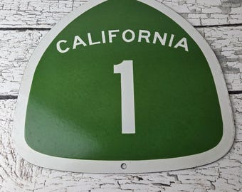 May include: A shield-shaped metal road sign with a green background and a white border. The text CALIFORNIA is printed in white at the top, with a large white number 1 centered below. There are two small circular mounting holes at the top and bottom.