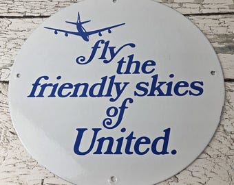 May include: A circular white metal sign featuring a blue graphic of an airplane and the blue text "fly the friendly skies of United." The sign has four small mounting holes positioned at the top, bottom, left, and right edges of the circular frame.