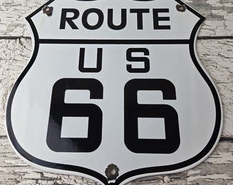 May include: A white metal shield-shaped sign with black borders and black lettering that reads ROUTE US 66. The sign features three small circular holes with visible rust around the edges, positioned at the top left, top right, and bottom center.
