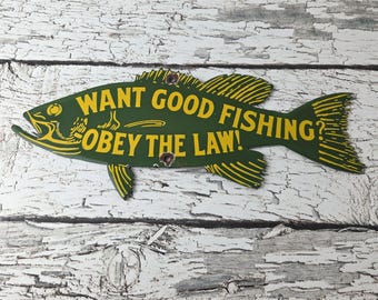 May include: A fish-shaped metal sign in dark green with yellow lettering that reads WANT GOOD FISHING? OBEY THE LAW! The sign features yellow fin and tail details, a pointed head, and two small circular holes for mounting purposes.
