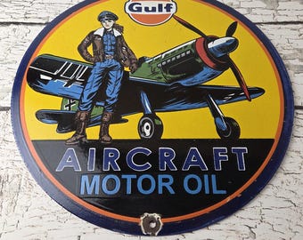 May include: Round metal sign with a yellow background featuring a vintage airplane and a person in a pilot's uniform. The sign has the Gulf logo and the words "AIRCRAFT MOTOR OIL". The sign is blue, black, red, and brown.