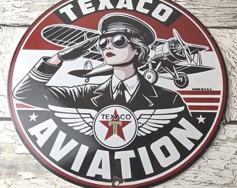May include: Round Texaco Aviation sign featuring a woman in a pilot's uniform saluting, with a biplane in the background. The sign is red, black, and white, with the Texaco logo and the word "AVIATION". Made in the U.S.A.