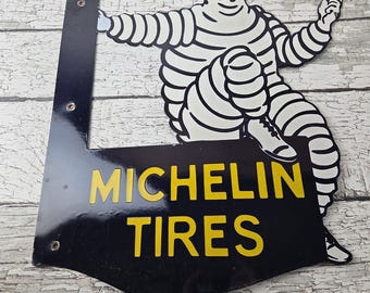 May include: Vintage Michelin Tires metal sign featuring the Michelin Man in white and black. The sign has a black background with yellow text that reads "MICHELIN TIRES". The sign is mounted on a black metal frame.