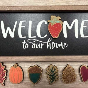 May include: A wooden sign with the text "WELCOME to our home" in white lettering on a black background.  Below the sign are seven wooden ornaments: a red and green leaf, an orange pumpkin, a brown and green acorn, a green and brown pine branch, a brown pine cone, a red heart, and a red strawberry.