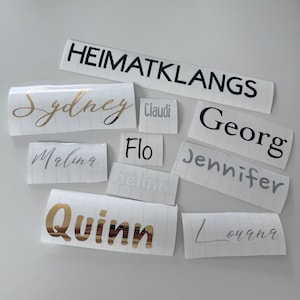 May include: A collection of white labels with various names written in black, gold, and silver lettering. The names include Sydney, Selina, Quinn, Louana, Marlin, Jennifer, Georg, Flo, Claudi, and Heimatklangs.