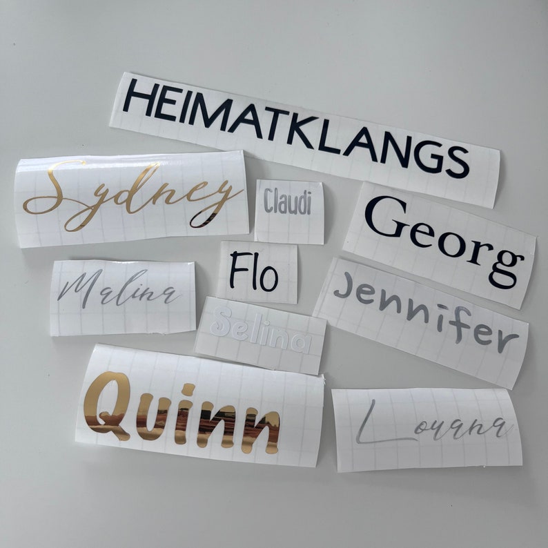 May include: A collection of white labels with various names written in black, gold, and silver lettering. The names include Sydney, Selina, Quinn, Louana, Marlin, Jennifer, Georg, Flo, Claudi, and Heimatklangs.
