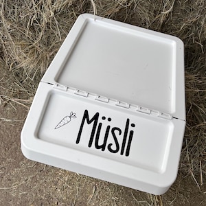 May include: White plastic pet food dish with a hinged lid. The dish has a black and white sticker that says "M&uuml;sli" with a small carrot graphic.
