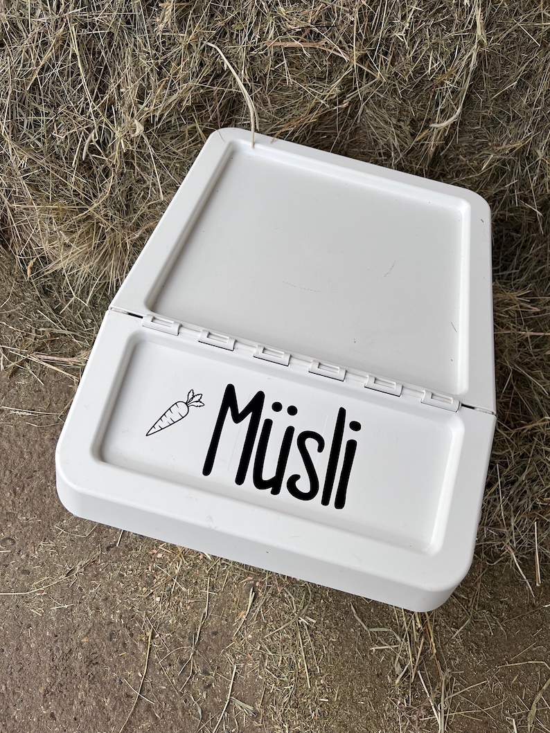 May include: White plastic pet food dish with a hinged lid. The dish has a black and white sticker that says "M&uuml;sli" with a small carrot graphic.