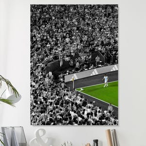 May include: A black and white photograph of a crowded stadium with a single soccer player in a light blue and white jersey standing on a vibrant green corner of the field. The stands are packed with spectators, creating a high-contrast visual.