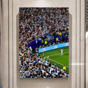 May include: A rectangular wall art print depicting a high-angle view of a crowded soccer stadium. A player in a white and light blue uniform stands on the green grass near the corner flag, while a large crowd fills the stands in the background.