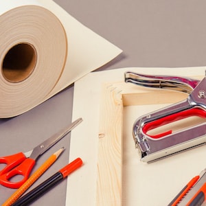 Op de afbeelding: Een canvas-spanraam met een nietpistool, een stanleymes, een rol canvas, een schaar en potloden. Het canvas is wit en het spanraam is van hout.