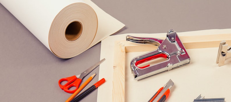 Op de afbeelding: Een canvas-spanraam met een nietpistool, een stanleymes, een rol canvas, een schaar en potloden. Het canvas is wit en het spanraam is van hout.