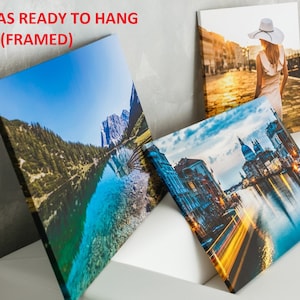 Op de afbeelding: Drie ingelijste canvas prints klaar om op te hangen. De eerste print toont een bergmeer met een blauwe lucht en groene bomen. De tweede print toont een stadsbeeld met een kanaal en gebouwen. De derde print toont een vrouw in een witte hoed en jurk die over straat loopt.