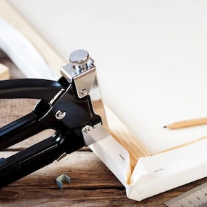 Op de afbeelding: Een zwarte canvas-spanner met een zilveren bovenkant en een zilveren nietjesverwijderaar. De nietpistool wordt gebruikt om een wit canvas aan een houten frame te bevestigen. Een potlood en een liniaal zijn ook zichtbaar in de afbeelding.