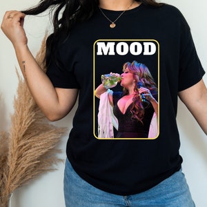 May include: Black t-shirt with a yellow rectangle featuring a woman drinking from a bottle and the word "MOOD" in white letters.