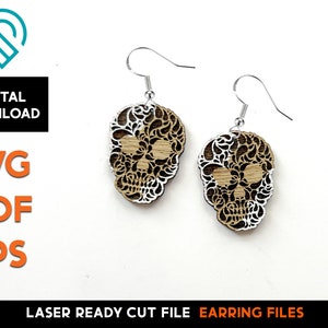 May include: A pair of laser cut wooden earrings shaped like a skull with intricate floral designs. The earrings are brown with white accents and have silver hooks.