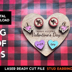 May include: A set of laser-cut wooden stud earrings featuring a heart with a dagger, a heart with the text "F*CK", a heart with the text "OFF", and two no-entry signs with a cupid inside. The earrings are displayed on a heart-shaped wooden base with the text "HAPPY FUCKING VALENTINE'S DAY".