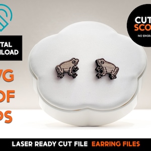 May include: A pair of laser cut wooden earrings featuring a frog design. The earrings are on a white display stand. The image includes the text "CUT & SCORE NO ENGRAVING".