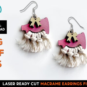 May include: A pair of laser cut wooden earrings featuring a pink megaphone with a gold bow on top. The earrings are accented with white macrame tassels.