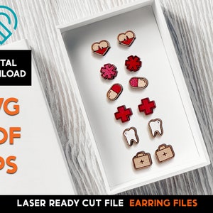 May include: A white box with a variety of laser cut wooden earring designs. The designs include hearts, stars, crosses, pills, teeth, and a first aid kit. The earrings are all red and white.
