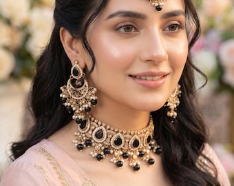 May include: A gold-toned jewelry set featuring a choker necklace, matching drop earrings, and a maang tikka. Each piece is adorned with black teardrop-shaped stones, clear crystals, and small white pearls arranged in a detailed filigree metal design.