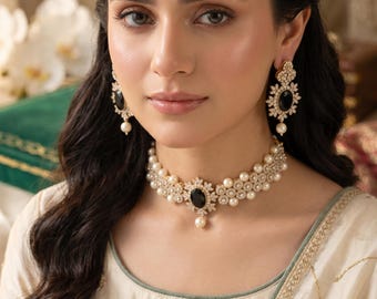 May include: A matching jewelry set featuring a necklace, earrings, and a headpiece. The set includes a black gemstone surrounded by a gold-colored filigree design, accented with pearls. The necklace and headpiece are connected by a strand of pearls.