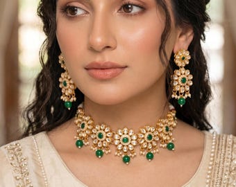 May include: A gold-toned jewelry set consisting of a choker necklace, drop earrings, and a maang tikka. Each piece features a floral design with white stones, a central green stone, and small green bead accents hanging from the bottom edges.