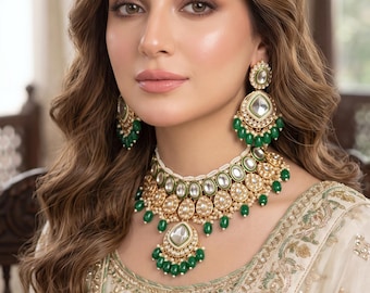 May include: A gold-toned jewelry set featuring a choker necklace, matching drop earrings, and a maang tikka. Each piece is adorned with white stones, small pearls, and dangling green beads, set against a cream-colored garment with gold embroidery.