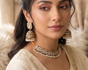 May include: A gold-toned jewelry set featuring a choker necklace, jhumka earrings, and a maang tikka. Each piece is crafted with clear stones and small white pearl accents. The necklace sits at the base of the neck, while the earrings are bell-shaped.